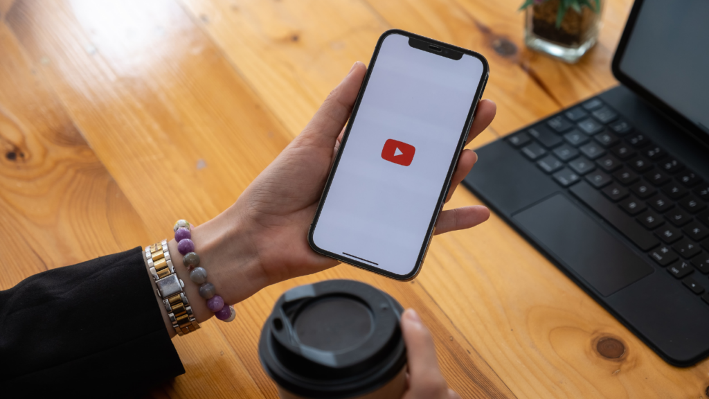 Person holding a smartphone with the YouTube logo on screen next to a laptop